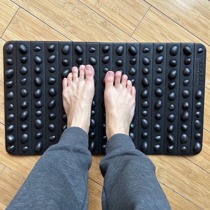 a top down view of a pair of bare feet standing on the Toe Spacer Rock Mat - a black foam mat with rock-shaped bumps for texture